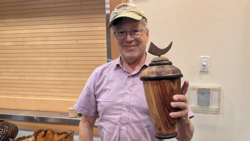 Fred Colman smiles while holding up a turned camphor wood vase topped with a crescent moon finial. He is wearing glasses, a pink checkered shirt, and a beige cap that reads "West Bay Woodturners," standing in front of a light wooden slat wall with other wood projects visible on a table to his left.
