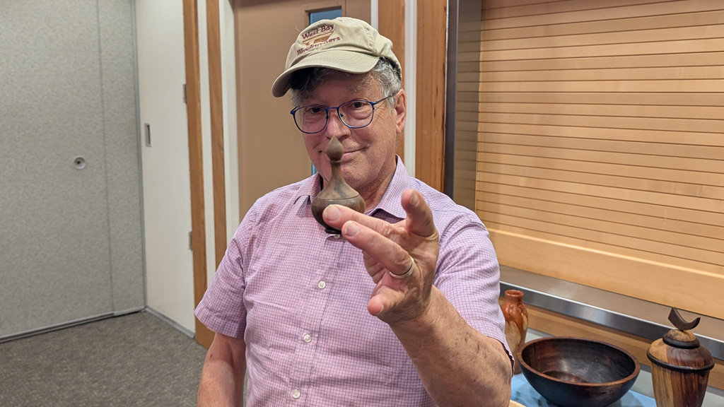 Fred Colman, wearing a beige “West Bay Woodturners” cap and wire-rimmed glasses, smiles as he holds up a miniature turned wooden box between his thumb and fingers. He is standing at a club meeting where other woodturning projects, including a dark bowl and a tall vase, are displayed on a table in the foreground, against a wall with horizontal wood slats.