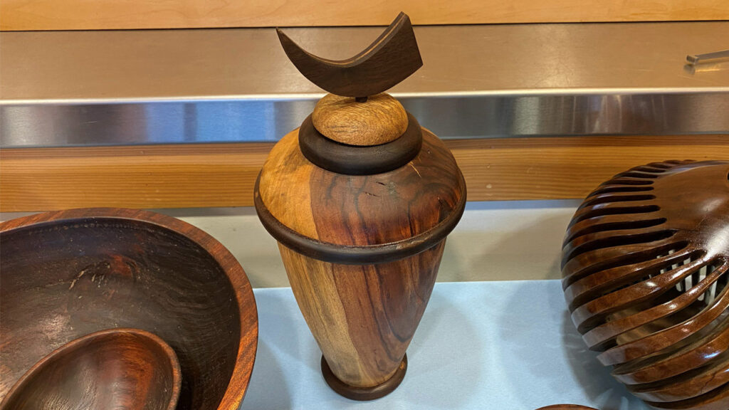 A wooden lidded vessel featuring a distinctive crescent-moon-shaped finial stands centrally on a light blue surface at the West Bay Woodturners meeting. The turned vase displays varied wood grain with light amber and deep brown tones, accented by dark rings around the lid rim and shoulder. To the left sits a large wooden bowl with a small ladle, while to the right stands an intricate, ribbed wooden sculpture.