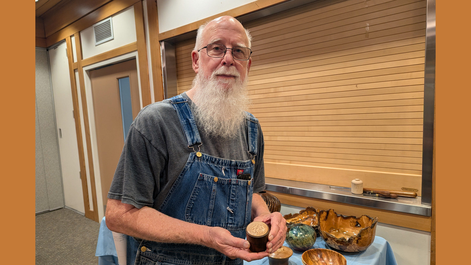 At a West Bay Woodturners club meeting, an older man, Harvey Klein, with a long white beard and glasses, wears denim overalls while holding up a small lidded wooden box. Several other turned wood pieces, including large natural-edge bowls and a glazed bowl, are displayed on a table behind him, set against light wood paneling.