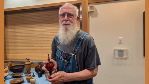 Harvey Klein, a man with a long white beard and glasses, wearing denim overalls, stands holding up a small, polished reddish-brown wooden vase. Behind him, a table displays an assortment of other woodturned projects, including bowls and vessels, at the West Bay Woodturners meeting.