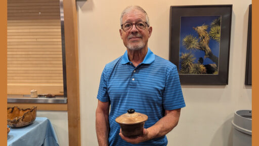 A smiling man, Jerri Galli, with short gray hair and glasses, stands indoors wearing a blue striped polo shirt. He holds a small, lidded wooden vessel in his hands. To his left, on a table covered with a light blue cloth and a nearby workbench, are other turned wood projects, including a large burl bowl and woodturning tools. Behind him hangs a framed photograph of a Joshua tree against a blue sky. The scene is from an exhibition at the West Bay Woodturners meeting, where members displayed their recent projects.