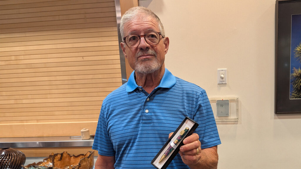 Jerry Galli, a man with grey hair and glasses wearing a blue striped polo shirt, holds up a black display box containing a handmade turned pen with brass hardware. He is pictured at a West Bay Woodturners meeting, standing in front of a wall with horizontal wood slats and other wooden projects visible on a counter to the left.