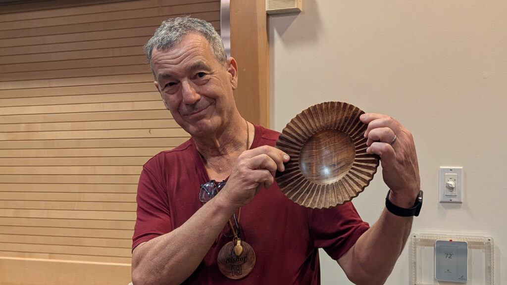 Jon Bishop smiles as he holds up a dark wooden bowl with a fluted, scalloped rim, showcasing a recent project at the West Bay Woodturners meeting. He is wearing a maroon T-shirt and has a large wooden medallion around his neck engraved with "Jon Bishop" and the "JB" monogram.