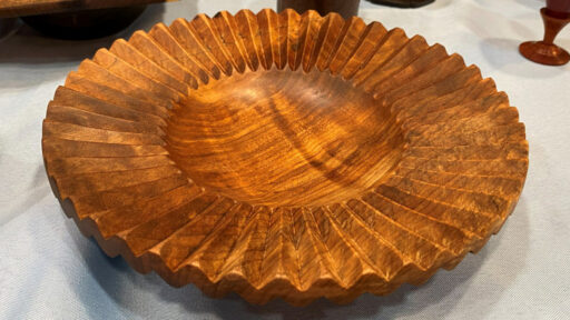 A hand-turned wooden platter by Jon Bishop featuring deep flutes that radiate outward from a smooth, concave center bowl to create a scalloped, sunburst-like rim. The wood has a rich, amber-brown tone with visible grain patterns highlighted by a glossy finish.