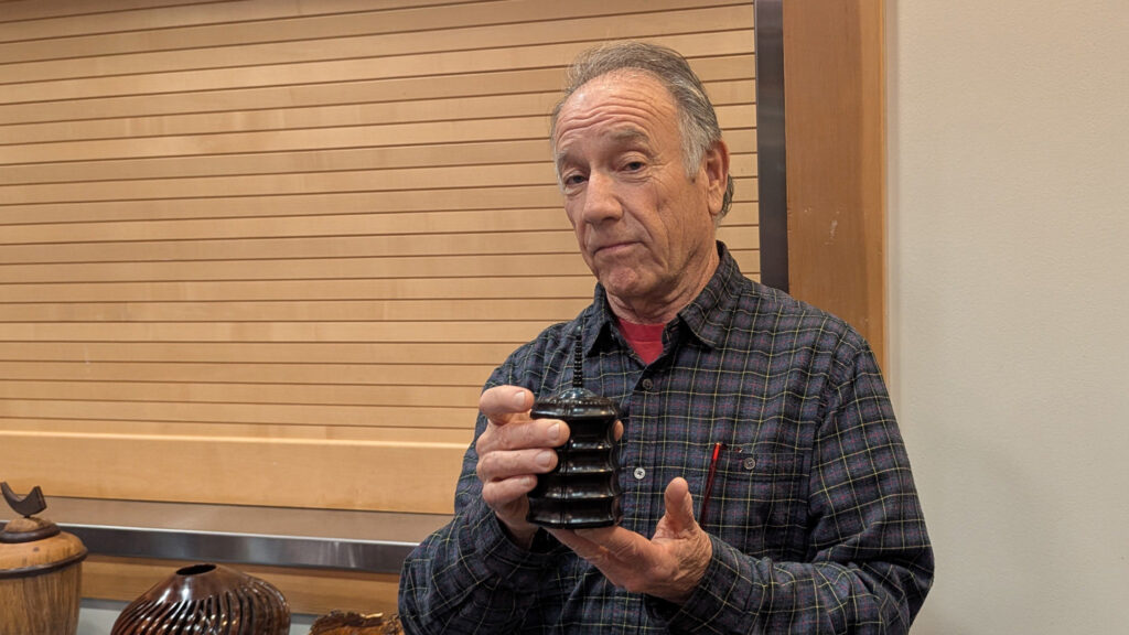 Jon Sauer, wearing a blue plaid button-down shirt over a red T-shirt, is holding and displaying a small, dark, wooden box that has a thin finial protruding from the top. He looks directly at the camera while supporting the object with both hands. In the lower-left foreground, partially out of frame, are other wood-turned pieces, including a light-colored lidded jar and a dark bowl with vertical grooves.