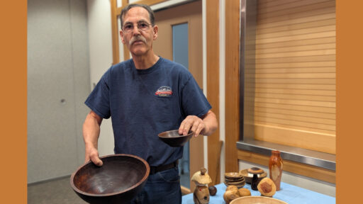At a West Bay Woodturners club meeting, Kelly Smith, a man with glasses and a mustache, stands behind a table displaying his recent woodturned projects. He holds a large, dark wooden bowl in his right hand and a smaller matching bowl in his left. On the light-blue tablecloth to his left sits an assortment of other turned-wood items, including stacked bowls, a vase, and decorative spheres.