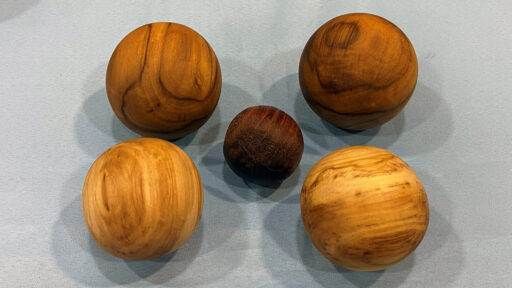 Five polished wooden spheres arranged on a light blue fabric background. Four larger spheres with golden and medium-brown wood grain surround a smaller, dark mahogany-colored sphere in the center. The image displays 'Turned Spheres' by Kelly Smith, shown at the West Bay Woodturners meeting.
