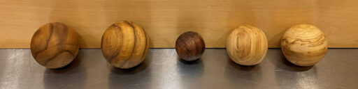 Five polished wooden spheres of varying sizes and wood species are lined up in a row on a brushed metal surface against a light-colored wooden backdrop. These appear to be woodturned croquet or Bocce balls.