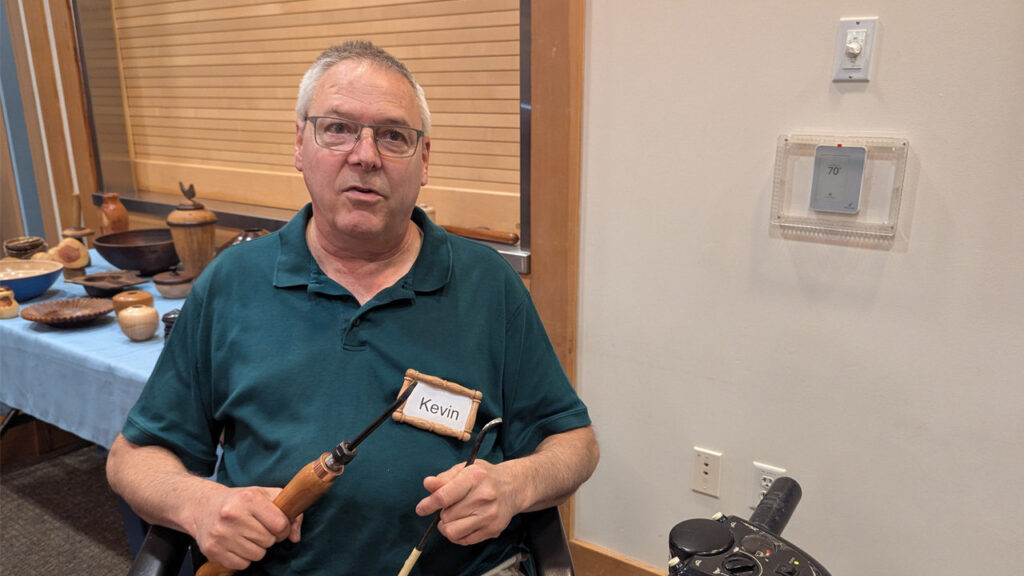 Kevin Lee holds two woodturning tools while near a table displaying turned wooden bowls and vessels at a West Bay Woodturners meeting. He wears glasses, a dark green polo shirt, and a name tag that reads "Kevin."