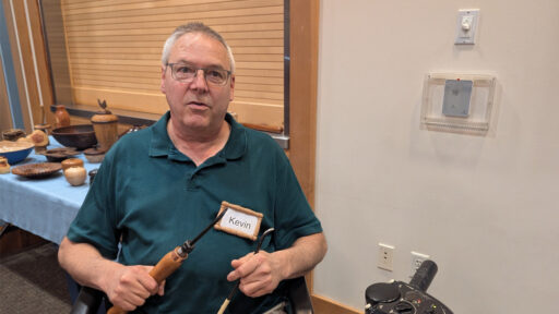 Kevin Lee holds two woodturning tools while near a table displaying turned wooden bowls and vessels at a West Bay Woodturners meeting. He wears glasses, a dark green polo shirt, and a name tag that reads "Kevin."