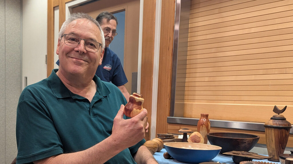 Kevin Lee, a smiling man with short grey hair and glasses, wearing a teal polo shirt, holds up a small, two-toned wooden lidded box for the camera. He turned it from juniper. To the right, a table displays an array of woodturning projects from a West Bay Woodturners meeting, including dark bowls, a bowl with a blue interior, and various turned vessels.