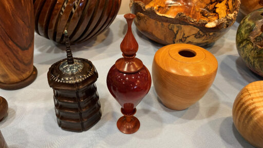 A collection of hand-turned wooden vessels and boxes displayed on a light-blue tablecloth. In the foreground, three distinct pieces are visible: a dark brown segmented box topped with an inlay and finial, a glossy reddish-brown lidded urn with an ornate stopper, and a smooth, light-orange wooden jar. Behind them are other woodturning projects, including a large natural-edge wooden bowl and a slatted wooden vase.
