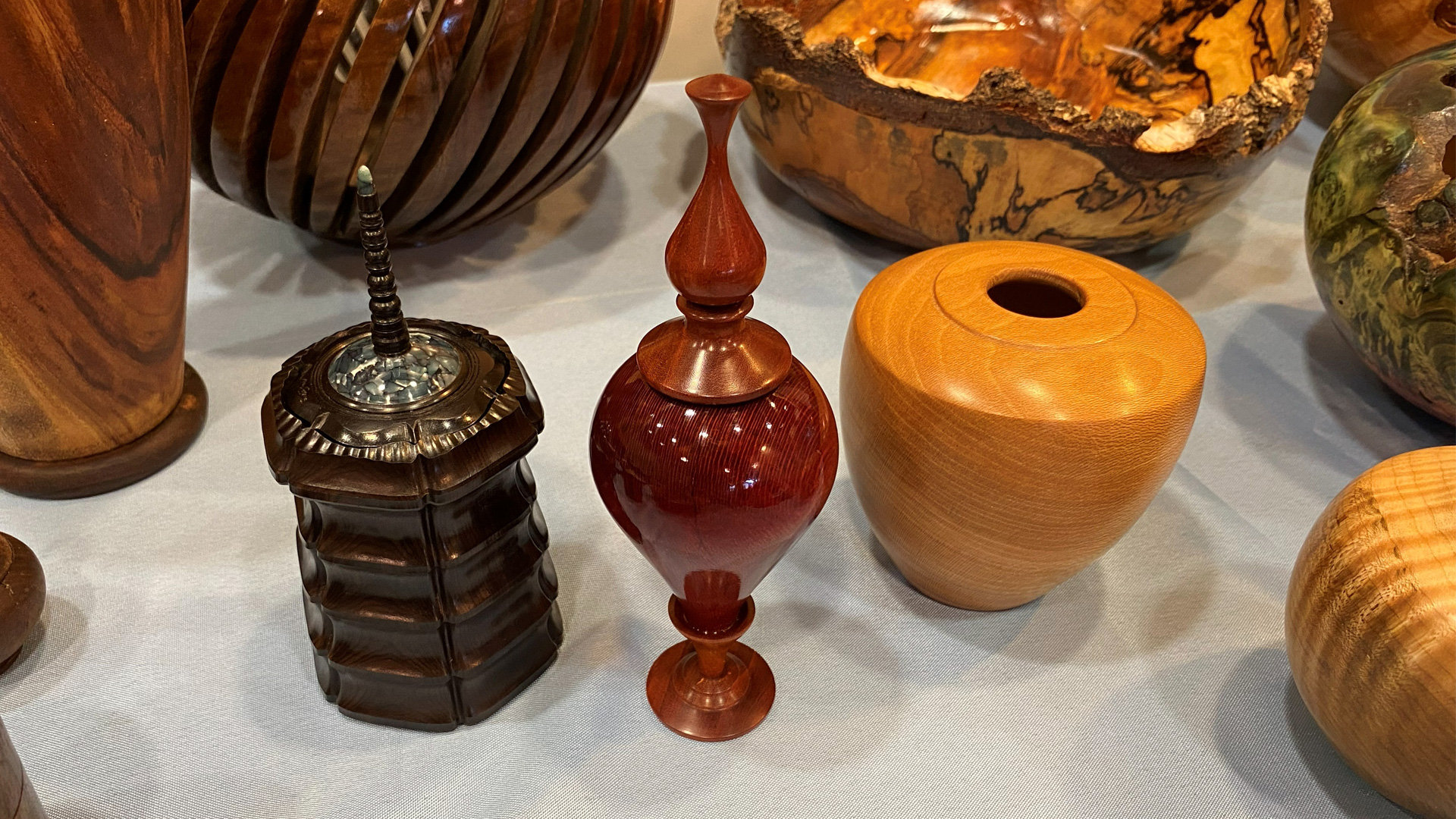 A collection of hand-turned wooden vessels and boxes displayed on a light-blue tablecloth. In the foreground, three distinct pieces are visible: a dark brown segmented box topped with an inlay and finial, a glossy reddish-brown lidded urn with an ornate stopper, and a smooth, light-orange wooden jar. Behind them are other woodturning projects, including a large natural-edge wooden bowl and a slatted wooden vase.