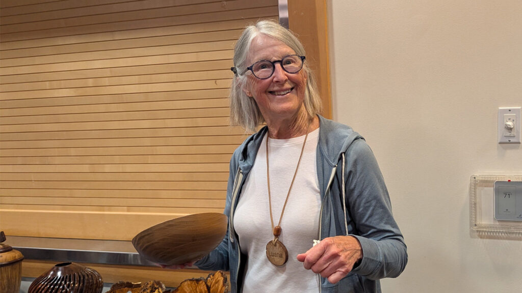 Luana Staiger smiles broadly while holding up a polished, brown turned wooden bowl at a West Bay Woodturners club meeting. She wears round glasses, a blue hooded sweatshirt, and a large wooden pendant necklace engraved with her name. In the foreground, other woodturning projects, including a dark vase and raw burl wood, are visible on a table against a light-colored slat wall.