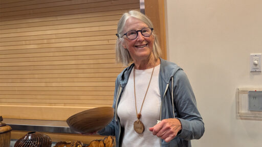 Luana Staiger smiles broadly while holding up a polished, brown turned wooden bowl at a West Bay Woodturners club meeting. She wears round glasses, a blue hooded sweatshirt, and a large wooden pendant necklace engraved with her name. In the foreground, other woodturning projects, including a dark vase and raw burl wood, are visible on a table against a light-colored slat wall.