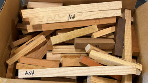 A top-down view of a cardboard box filled with rectangular wooden blanks for woodturning. The pieces vary in color from pale cream to dark reddish-brown, and several light-colored strips at the top and bottom are labeled "ASH" in black marker. These are pen blanks given out as participation prizes.