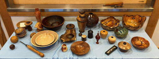A wide variety of woodturned items are displayed on a table covered with a light blue cloth at a West Bay Woodturners club meeting. The collection features an assortment of wooden bowls in different sizes and finishes—ranging from dark, polished woods to light maple—including large live-edge burl bowls on the right side and a nested set of light-colored bowls in the front left. Interspersed among the bowls are various turned objects, including tall vases, lidded jars, small finials, spheres, and a tiered black pagoda-like object. The show-and-tell display was arranged for the WBW meeting and the woodturners newsletter.