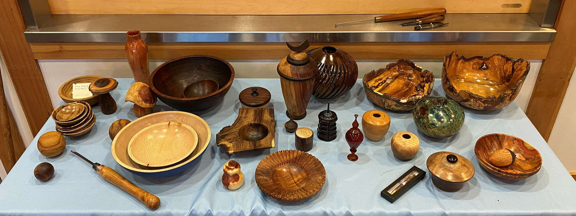 A wide variety of woodturned items are displayed on a table covered with a light blue cloth at a West Bay Woodturners club meeting. The collection features an assortment of wooden bowls in different sizes and finishes—ranging from dark, polished woods to light maple—including large live-edge burl bowls on the right side and a nested set of light-colored bowls in the front left. Interspersed among the bowls are various turned objects, including tall vases, lidded jars, small finials, spheres, and a tiered black pagoda-like object. The show-and-tell display was arranged for the WBW meeting and the woodturners newsletter.