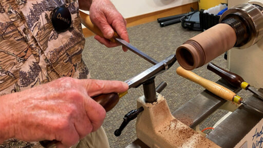 A close-up photo captures a woodturning demonstration at the West Bay Woodturners meeting. A man wearing a name tag that reads "Dennis" is using a technique called thread chasing on a lathe. He uses a supporting tool to hold a thread-chasing tool steady against the spinning wood, cutting deep spiral threads into the end of a light-colored wooden box. Two other turning tools with wooden handles rest on the lathe bed nearby.