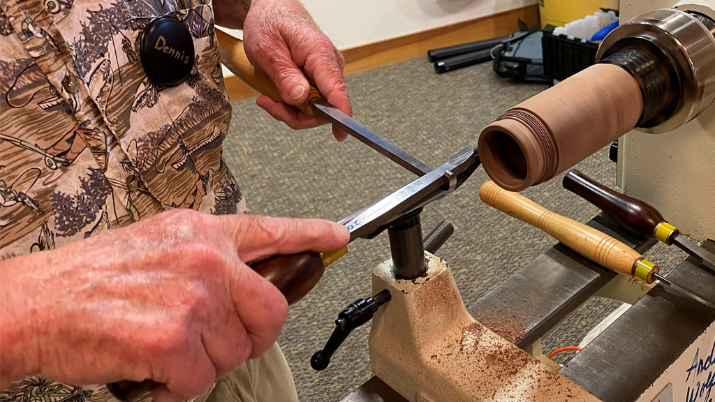 A close-up photo captures a woodturning demonstration at the West Bay Woodturners meeting. A man wearing a name tag that reads "Dennis" is using a technique called thread chasing on a lathe. He uses a supporting tool to hold a thread-chasing tool steady against the spinning wood, cutting deep spiral threads into the end of a light-colored wooden box. Two other turning tools with wooden handles rest on the lathe bed nearby.