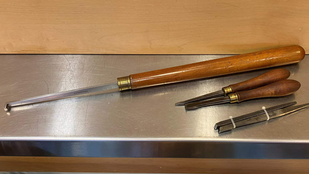 A collection of specialized woodturning thread chasing tools is arranged on a brushed stainless-steel surface against a light-colored wooden board. The largest tool features a long, polished wooden handle with a brass ferrule and a slender metal blade that ends in an upward-curving hook. Beside it lie two smaller threading scrapers with dark wooden handles and brass collars. Below them is a pair of metal thread-chasing inserts with serrated edges, held closed by a white rubber band.