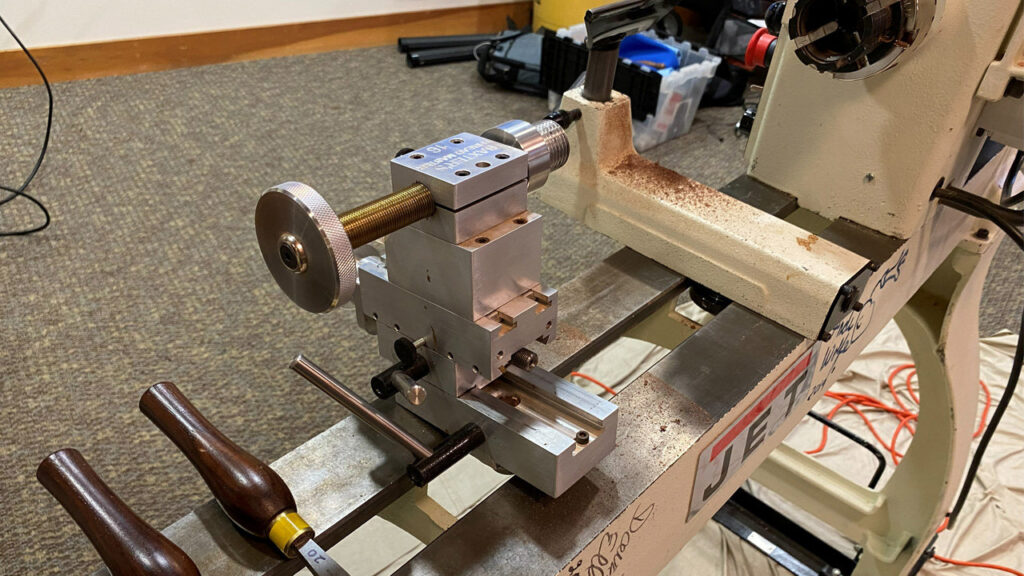 A close-up view of a Baxter threading jig mounted on a JET wood lathe. The device features a large brass lead screw with a knurled adjustment wheel for precise movement and is constructed from stacked aluminum blocks. Wooden-handled turning tools rest nearby on the lathe bed, and the background shows a carpeted room, indicating this was displayed at a local woodturning club meeting.