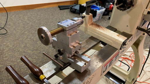 A close-up view of a Baxter threading jig mounted on a JET wood lathe. The device features a large brass lead screw with a knurled adjustment wheel for precise movement and is constructed from stacked aluminum blocks. Wooden-handled turning tools rest nearby on the lathe bed, and the background shows a carpeted room, indicating this was displayed at a local woodturning club meeting.