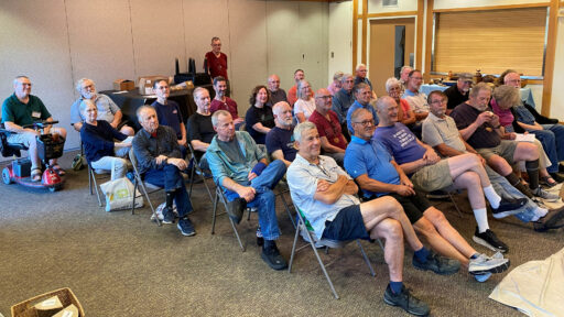 A group of people, mostly men dressed in casual clothing, are seated in rows of folding chairs facing toward the right at the West Bay Woodturners club meeting. They are an audience watching a thread chasing woodturning demonstration by Dennis Lillis.