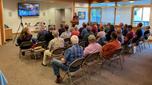This image captures an indoor meeting of the West Bay Woodturners club. Viewed from behind the audience, rows of attendees sit in folding chairs facing a speaker standing at a wooden podium near the back wall. To the left, a large wood lathe sits against the wall beneath a mounted television screen that displays a video feed, while a camera on a tripod is set up nearby. The room features light wood trim and large windows on the right side that look out onto a deck.