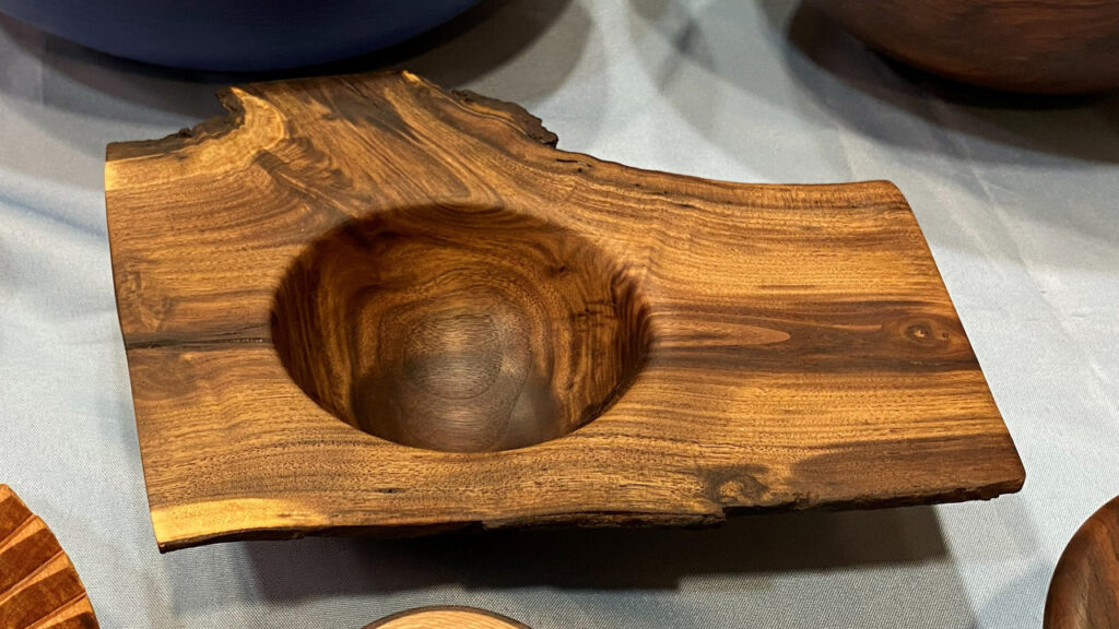 A handcrafted wooden vessel known as a "winged bowl," featuring a rectangular slab of wood with natural live edges and a deep, circular depression turned into the center. The wood displays rich golden-brown tones with distinct dark striping patterns. It sits on a light blue tablecloth, displayed at the West Bay Woodturners meeting alongside other club members' projects.