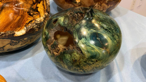 A close-up view of a hand-turned wooden hollow form made from burl, featuring a glossy finish dyed in swirling shades of deep green and teal. The bowl has a rough, natural, dark-bark rim and sits on a light blue cloth next to another turned bowl with warm amber and orange tones.