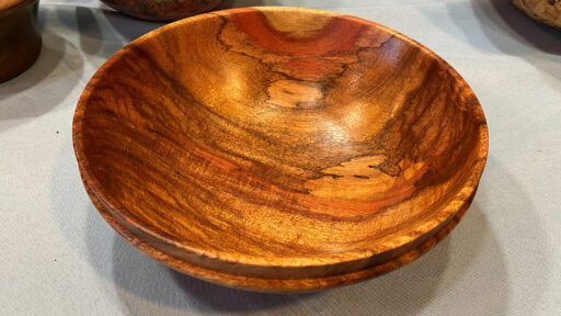 A polished, turned wooden bowl by Dean Caudle, featuring rich amber and reddish-brown grain patterns with dark streaks, displayed on a cloth surface at the West Bay Woodturners meeting.