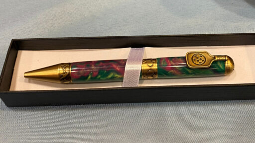 A custom turned pen by Jerri Galli rests horizontally in a long black presentation box lined with white fabric. The barrel features a vibrant, swirled pattern of translucent resin in shades of green, deep pink, and yellow, echoing pickleball court colors. Brass hardware accents include a pointed cone tip, a decorative middle band carved with pickleball motifs (holes or paddles), and a unique brass clip at the cap end that resembles a small slotted paddle. A white satin ribbon wraps around the center to secure the pen in place.