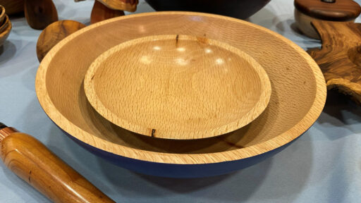 Two hand-turned oak bowls nested together, displayed on a table at the West Bay Woodturners meeting. The larger bowl has a wide rim and a glimpse of blue paint on its exterior base, while the smaller inner bowl showcases light honey-colored wood grain with small natural imperfections. Other turned wooden objects, including a tool handle, surround the bowls in the background.