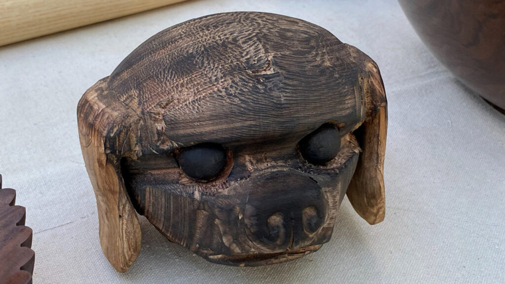 A wooden multi-axis turned sculpture of a dog's head with dark inset eyes and lighter-colored floppy ears by Angela Gunn.