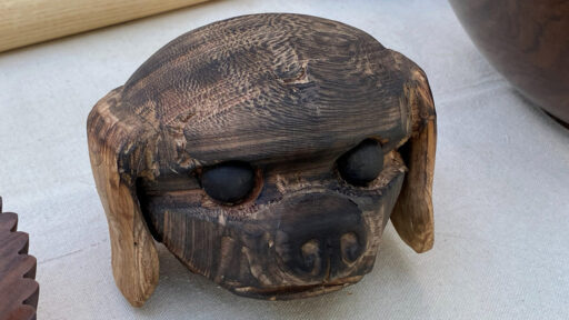 A wooden multi-axis turned sculpture of a dog's head with dark inset eyes and lighter-colored floppy ears by Angela Gunn.