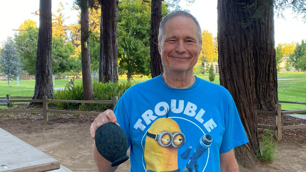 David Vannier, wearing a blue Minion T-shirt, holds a dark, textured wood-turned vase at the woodturning club picnic in Cuesta Park in 2026.