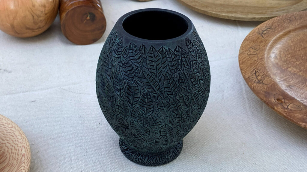 A dark, turned wooden vessel featuring a detailed carving of overlapping leaves covering its entire surface. Turned and carved by David Vannier.