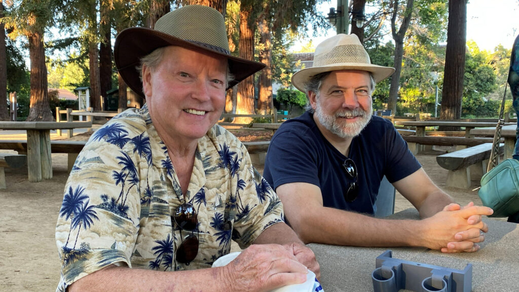 Dennis Lillis (left) and James Craig (right) sit at a picnic table outdoors, smiling at the camera. Dennis wears a brown wide-brimmed hat and a palm-tree-print shirt, while James wears a straw hat and a dark blue T-shirt.