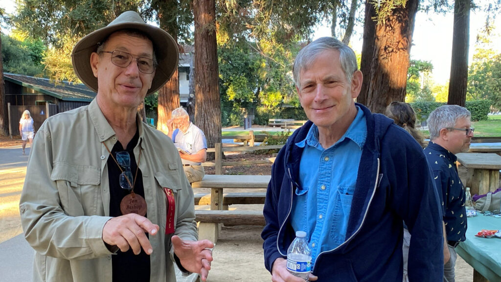Jon Bishop (left) and Tom Mandle (right) pose for a photo at the joint Silicon Valley Woodturners and West Bay Woodturners club picnic in Cuesta Park. Jon wears a hat and a wooden name pendant, while Tom holds a water bottle.