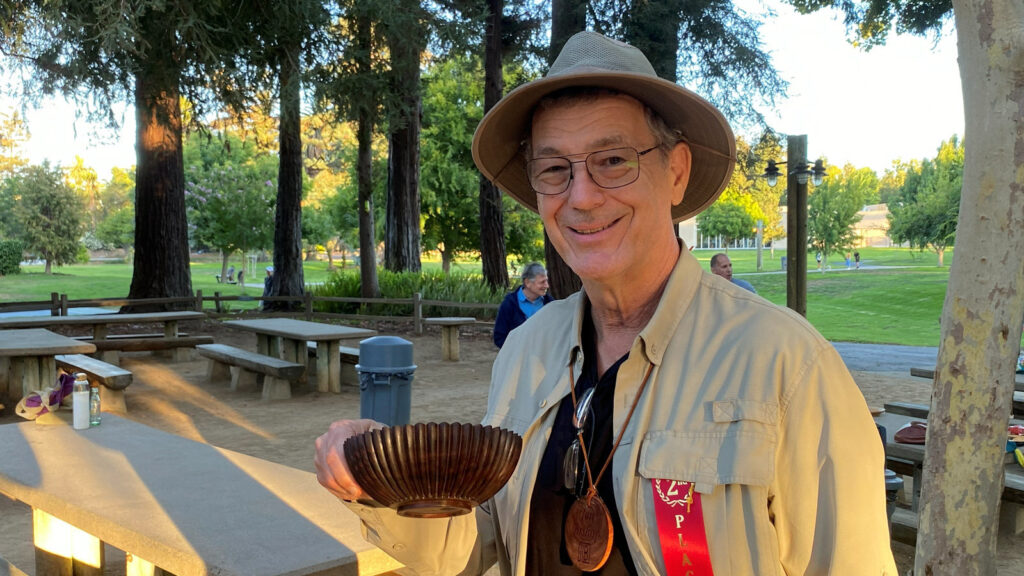 Jon Bishop holds a dark, ribbed wooden bowl at a woodturning club picnic, wearing a wooden name badge and a red 2nd place ribbon.