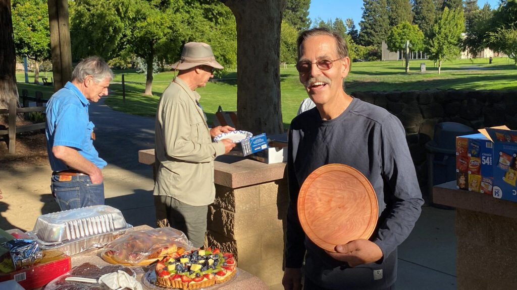At a joint picnic for the Silicon Valley Woodturners and West Bay Woodturners in Cuesta Park, Kelly Smith, a smiling man with a mustache, holds up a round wooden plate. In the background, other members gather around a table laden with food and dessert.
