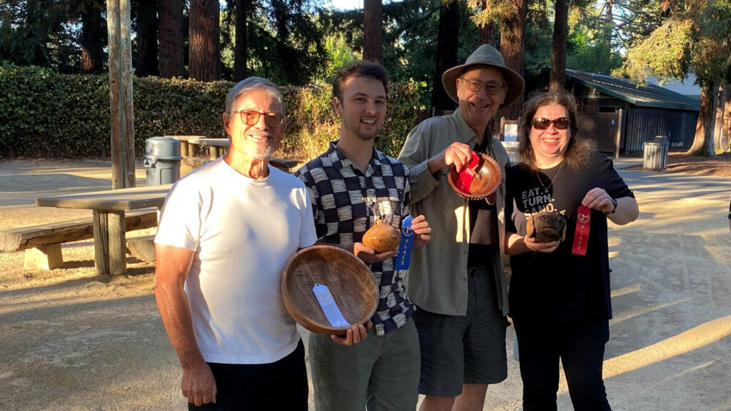 Four smiling people stand outdoors at a picnic table in Cuesta Park, holding wooden turned bowls and ribbons, celebrating their People's Choice award wins at a 2026 joint woodturning club picnic: Jerry Galli, Dylan McVoy, Jon Bishop, and Angela Gunn.