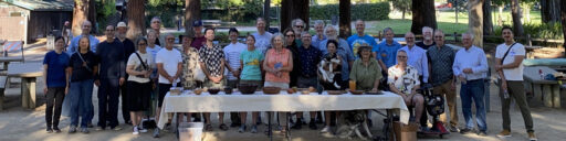 A group photo of members from the Silicon Valley Woodturners and West Bay Woodturners clubs gathered for a joint picnic at Cuesta Park, standing behind a table displaying various woodturning projects.