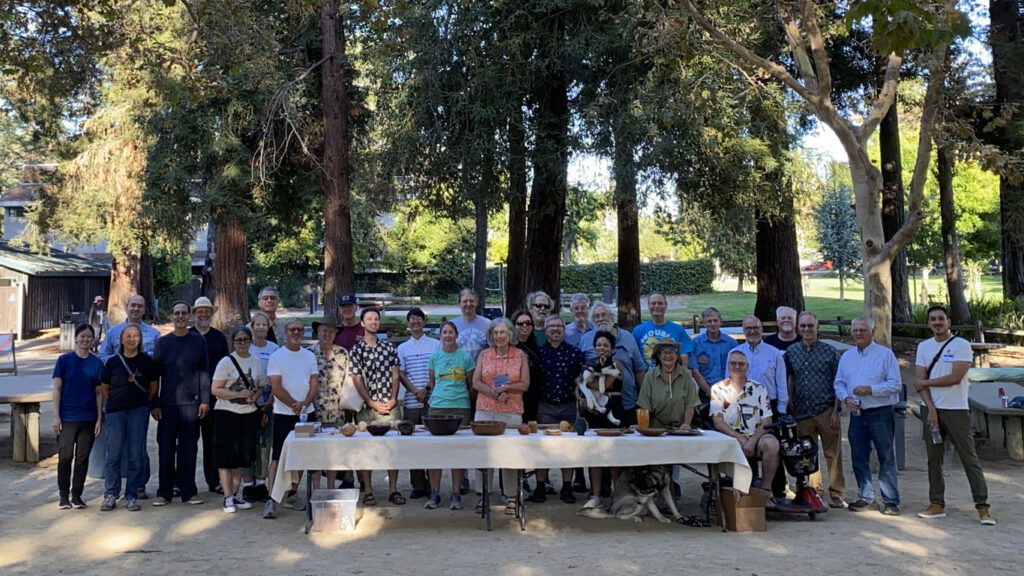 Group photo of members from the Silicon Valley Woodturners and West Bay Woodturners clubs posing behind a table displaying wooden bowls and turned projects at their joint picnic in Cuesta Park.