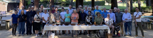 Group photo of members from the Silicon Valley Woodturners and West Bay Woodturners clubs at their joint picnic in Cuesta Park, Mountain View, CA, on August 5, 2026. The group stands behind a table displaying various woodturning projects, including wooden bowls, with dogs present.