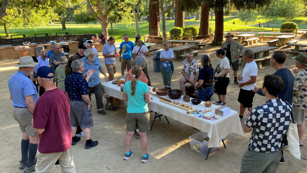 Members of the Silicon Valley Woodturners and West Bay Woodturners clubs gather around tables displaying woodturning projects, including bowls and tools, during their joint picnic at Cuesta Park in Mountain View, CA, on August 5, 2026.