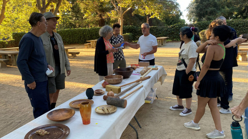 A group of woodturning enthusiasts gathers around a table displaying wooden bowls, mallets, and vases, chatting at an outdoor picnic.
