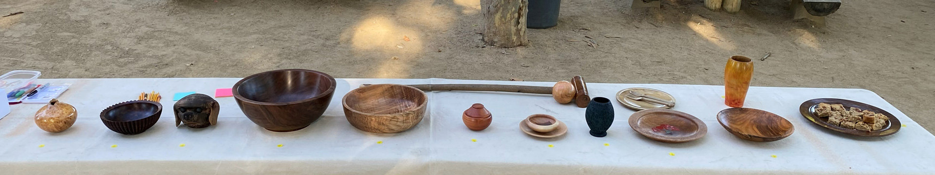 A long table covered in a white cloth displays a variety of woodturned items, including a large dark bowl, a light wood bowl, a carved dog head, a mallet, a small lidded box, a dark vase, plates, and a tall amber vase. A platter with food sits on the far right.